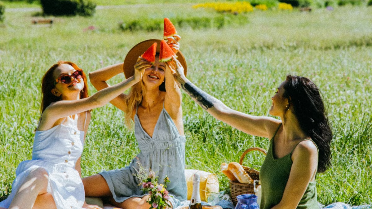 Three women enjoying a fun summer picnic with watermelon toast in a lush park setting.